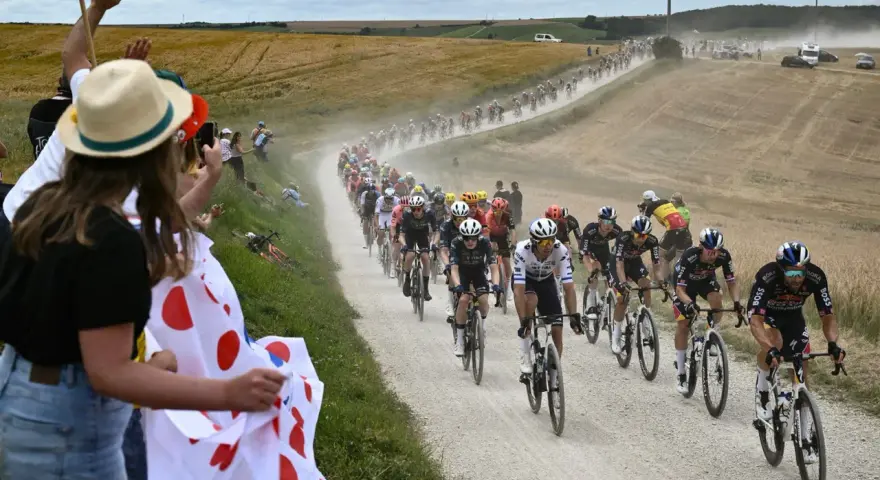 Spectators cheer as a peloton races on a dusty road during the Tour de France watch. Cyclists in colorful jerseys kick up dust in a dramatic scene.