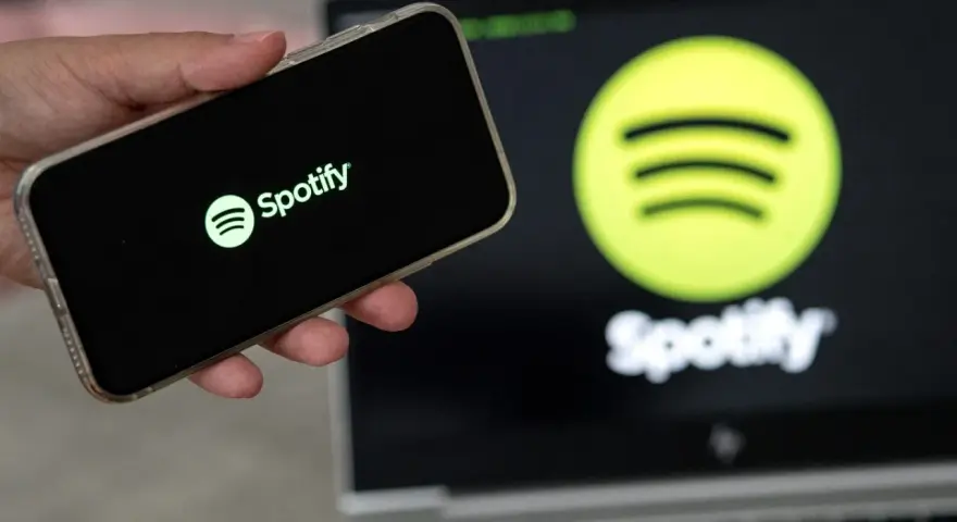 Dłoń trzyma smartfon z logo Spotify. W tle ekran laptopa z tym samym logo.