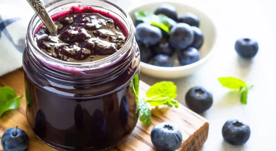 Vaso di marmellata di mirtilli fatto in casa, con mirtilli freschi e foglie di menta su un tagliere di legno.