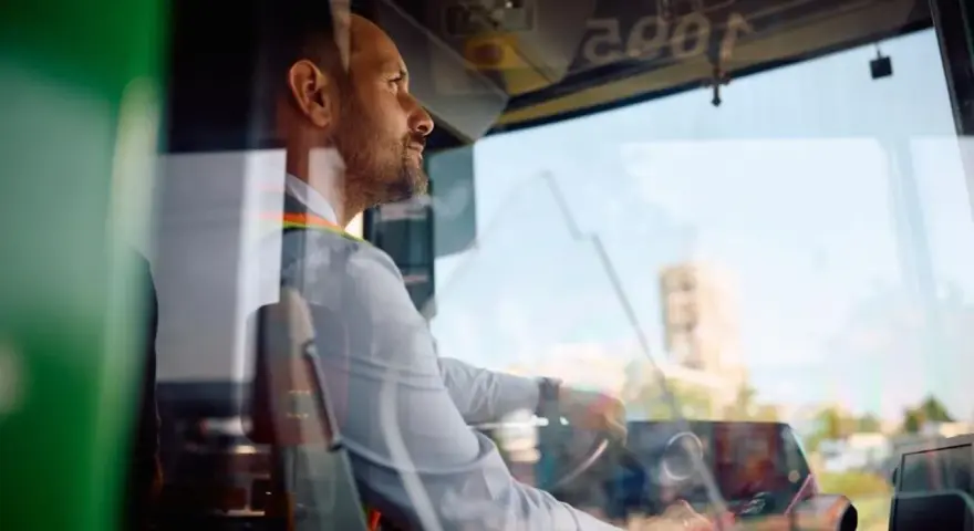 Skupiony kierowca autobusu prowadzi pojazd, trzymając kierownicę. Widać jego profil i fragment deski rozdzielczej.