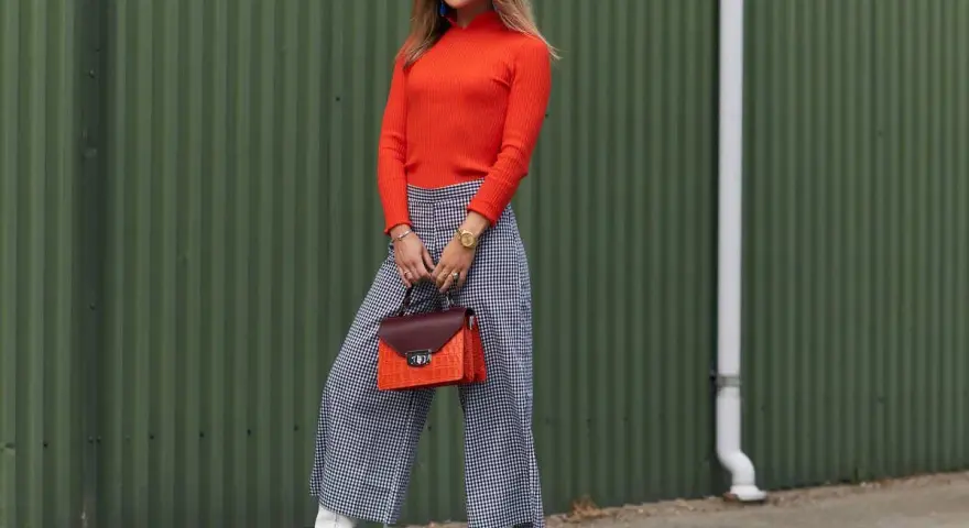 Kobieta w pomarańczowym golfie i kraciastych kulotach prezentuje stylową stylizację. Idealne buty do kulotów to białe botki na obcasie.