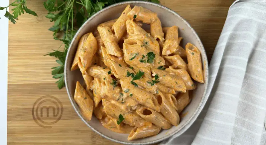 Penne alla vodka cremose e invitanti, guarnite con prezzemolo fresco, servite in una ciotola su un tagliere di legno con il logo MasterChef.