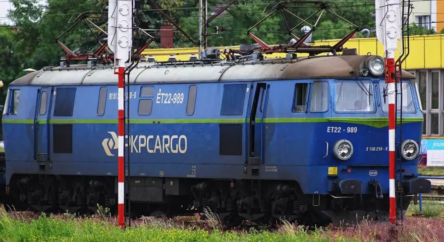 Niebieska lokomotywa PKP Cargo ET22-989 na tle zieleni i budynków.