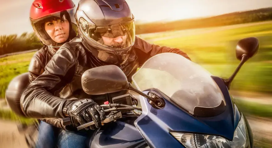 Coppia in moto, con lui alla guida e lei dietro, entrambi con caschi e giacche di pelle. Un'immagine che evoca la libertà di come guidare una moto.