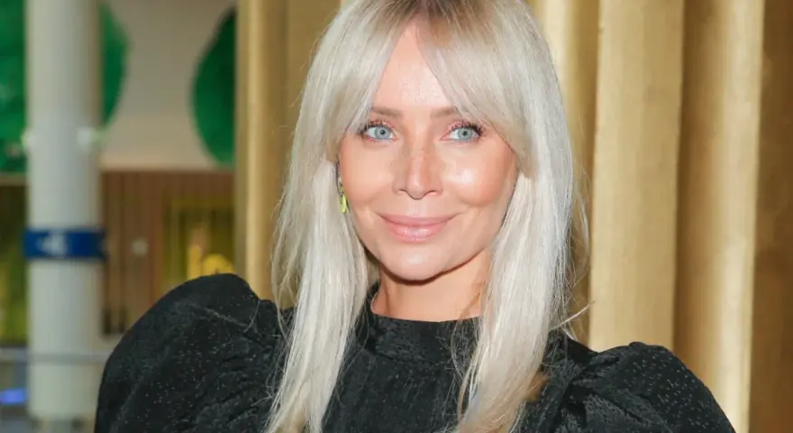 Blondynka z niebieskimi oczami i uroczą grzywką na boki, uśmiecha się lekko.