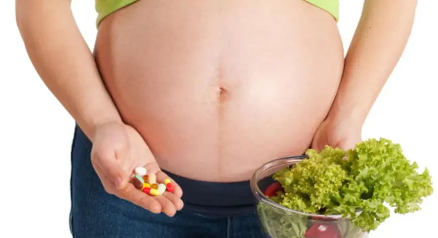 Kobieta w ciąży trzyma garść tabletek i miskę sałatki. Ważne jest żelazo dla kobiet w ciąży, czy wybrać suplementy czy zdrową dietę?