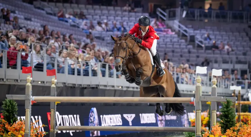 Cavalier en tenue rouge saute un obstacle lors d'une compétition équestre, visant la victoire à la **equestrian cup**.