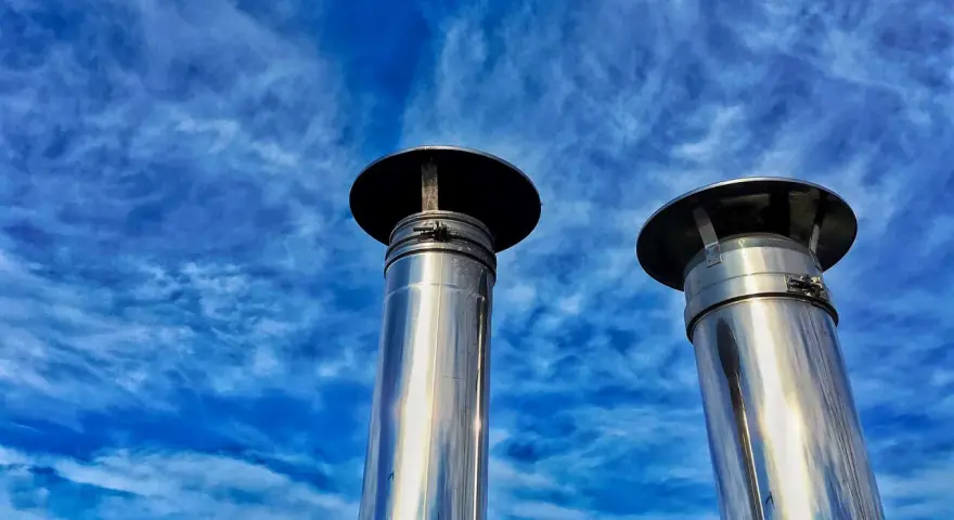 Due camini in acciaio inox, modello uni 7129 scarico a parete, brillano sotto un cielo blu con nuvole sottili.