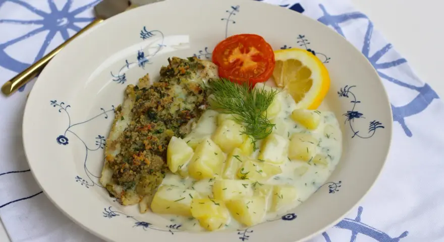En tallrik med panerad fisk, dillstuvad potatis, en tomatklyfta och en citronklyfta. Allt serveras på en tallrik med blått mönster.