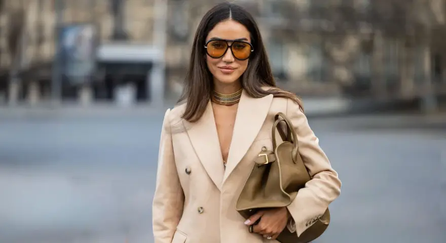 Mujer con gafas naranjas y blazer camel. Su bolso marrón y gargantilla dorada complementan el look, creando un estilo chic para combinar camel y azul.