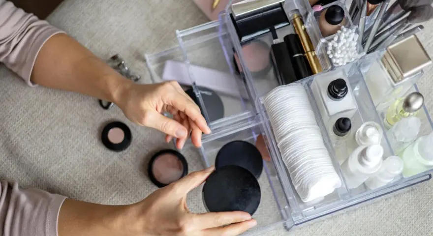 Hände sortieren Kosmetik in einem transparenten Organizer mit Pinseln, Puderdosen und Cremes.