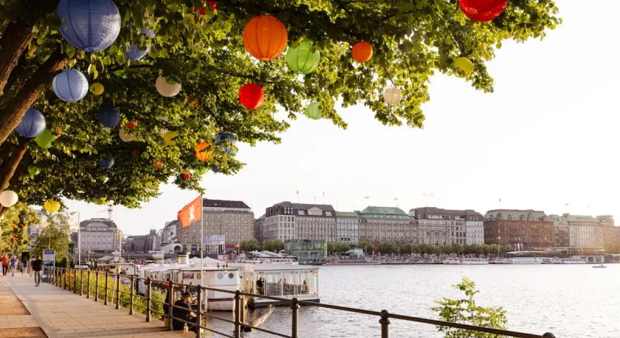 Bunte Lampions schmücken einen Baum am Wasser. Im Hintergrund die elegante Skyline von Hamburg mit Gebäuden und Booten.