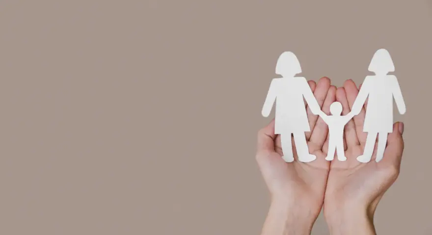 Dwie dłonie trzymają białe wycinanki: dwie postacie dorosłych i między nimi tęczowe dziecko.