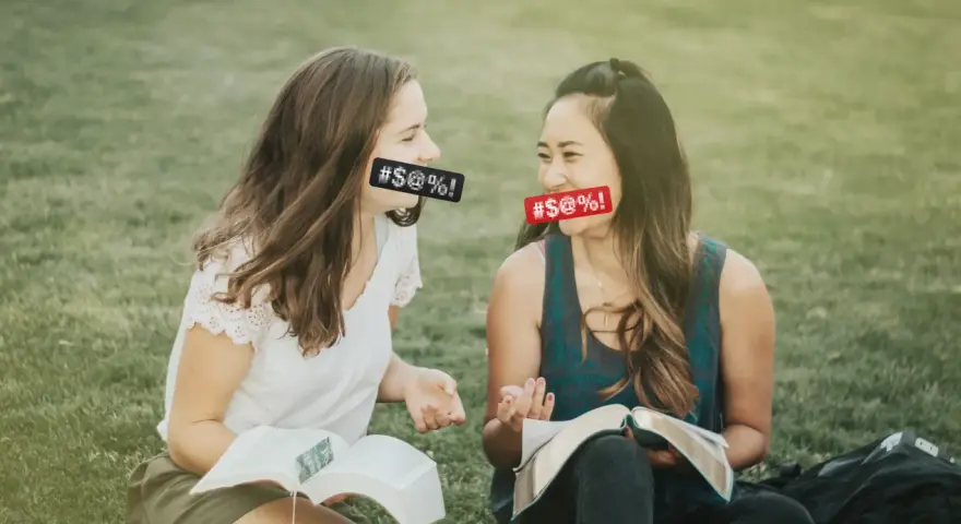 Two women with censored mouths discuss if cursing is a sin while reading books on the grass.