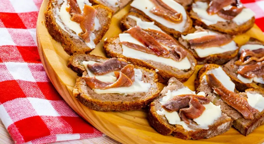 Na drewnianej desce ułożone są kromki chleba z masłem i anchois. To pyszne, proste przekąski.