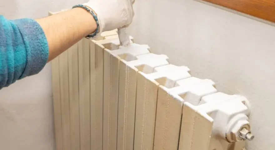 Mano con guanto bianco che dipinge un termosifone beige con vernice bianca, pronto per imbiancare la stanza.
