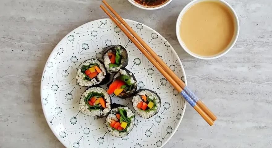 Sześć wegetariańskich rolek sushi z kolorowymi warzywami, przygotowanych z użyciem glonu do sushi, leży na talerzu obok sosów i pałeczek.