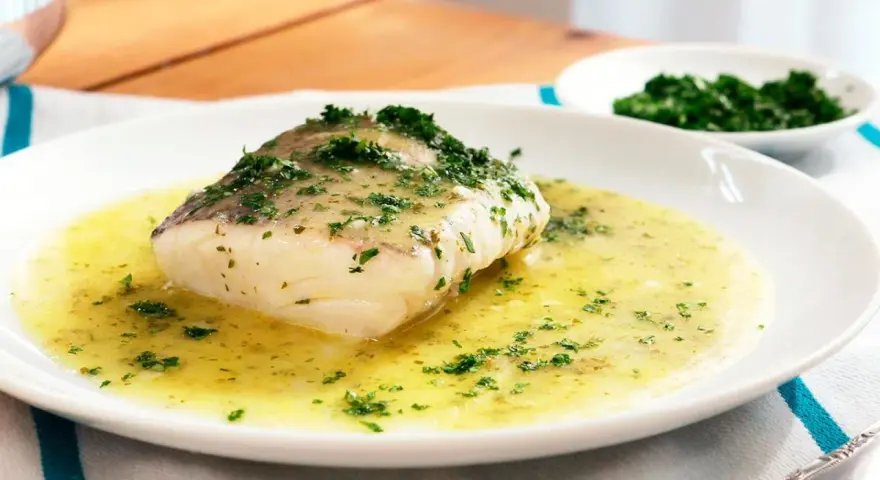 Un filete de merluza en salsa verde, con perejil picado, servido en un plato blanco.