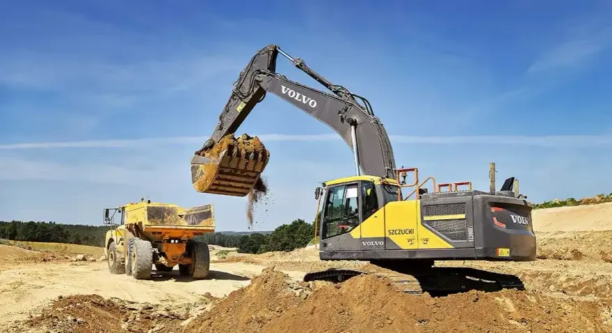 Koparka Volvo wysypuje ziemię do wywrotki. Prace ziemne, ułożenie rur w wykopie cennik bez materiału.