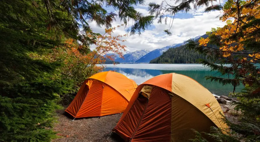 Två orangea tält står uppställda vid en sjö, omgivna av höstfärgad skog och berg. En perfekt plats för kamlunge camping.