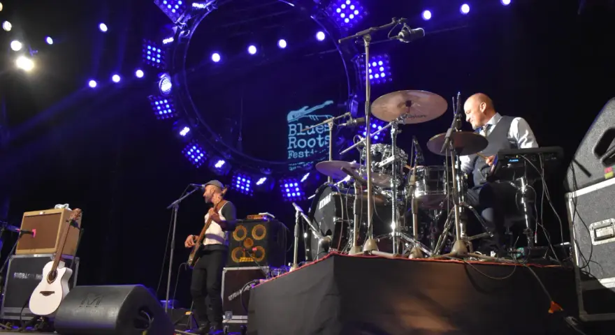 Un groupe joue du blues musique sous un grand cercle lumineux. Un bassiste à gauche, un batteur à droite.