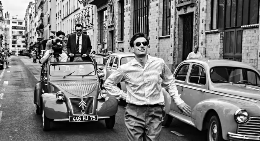 Un uomo con sigaretta in bocca, occhiali da sole e camicia a righe, in strada con auto d'epoca, evoca lo spirito de **la nouvelle vague**.