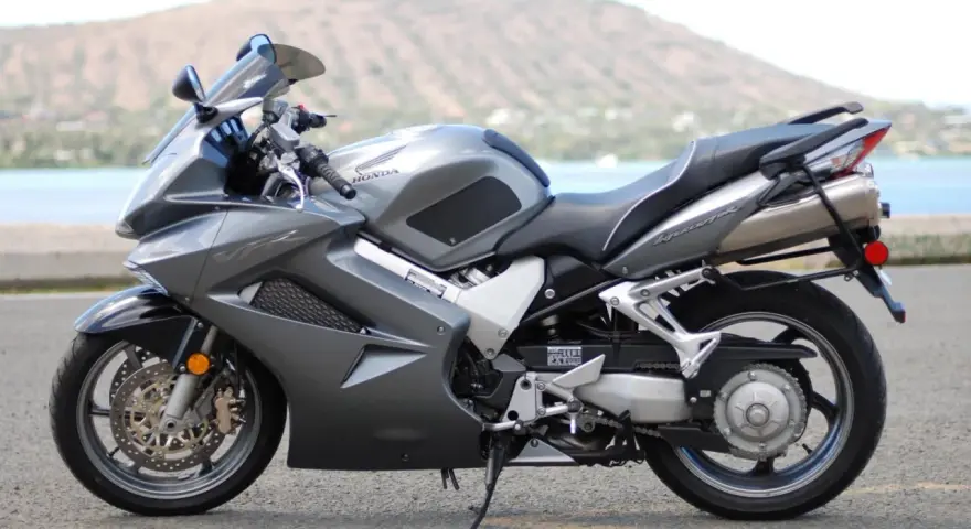 Szary motocykl Honda VFR 800 z widokiem na wodę i wzgórze w tle.