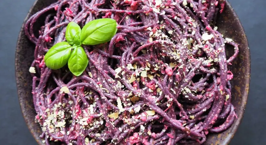 Fioletowy makaron spaghetti z buraczanym pesto, posypany orzechami i udekorowany listkiem bazylii.