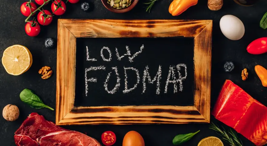 Na tablicy napis "LOW FODMAP". Wokół świeże produkty: mięso, ryba, pomidorki, cytryna, jajko, papryka, orzechy, jagody, szpinak.
