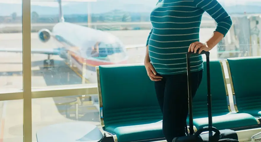 Ciężarna kobieta z walizką czeka na lotnisku. W tle widać samolot i pasażerskie fotele. Latanie samolotem w ciąży to wyzwanie, ale podróż dobiega końca.