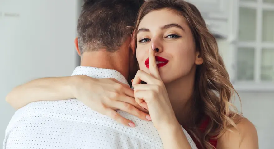 Une femme aux lèvres rouges fait signe de se taire en serrant un homme dans ses bras. Elle sait comment chauffer un mec.