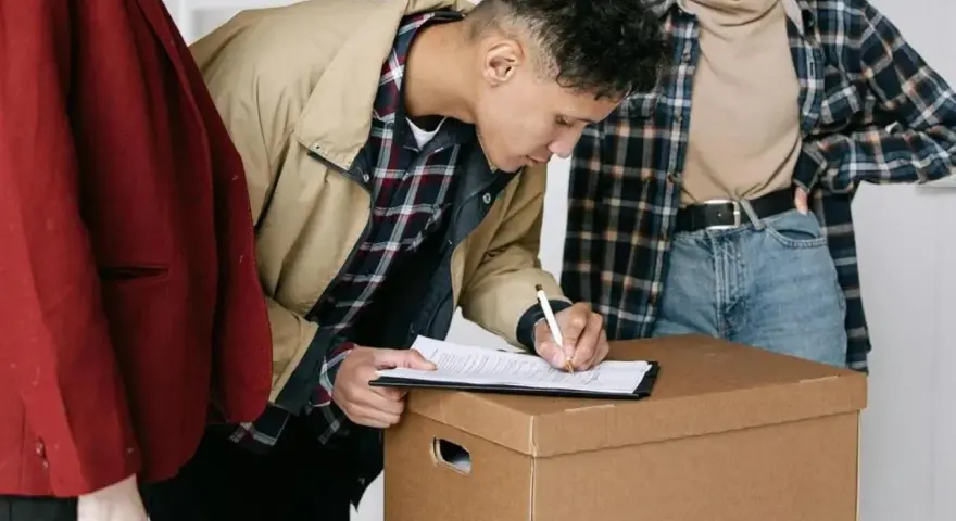 Joven firma contratos de alquiler sobre una caja de mudanza, acompañado de dos personas.