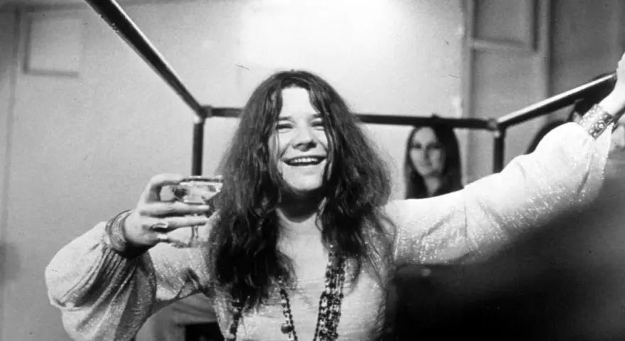 Janis Joplin z kieliszkiem w dłoni, uśmiechnięta. Jej charakterystyczne włosy i naszyjniki przywodzą na myśl jej niezapomniane utwory.