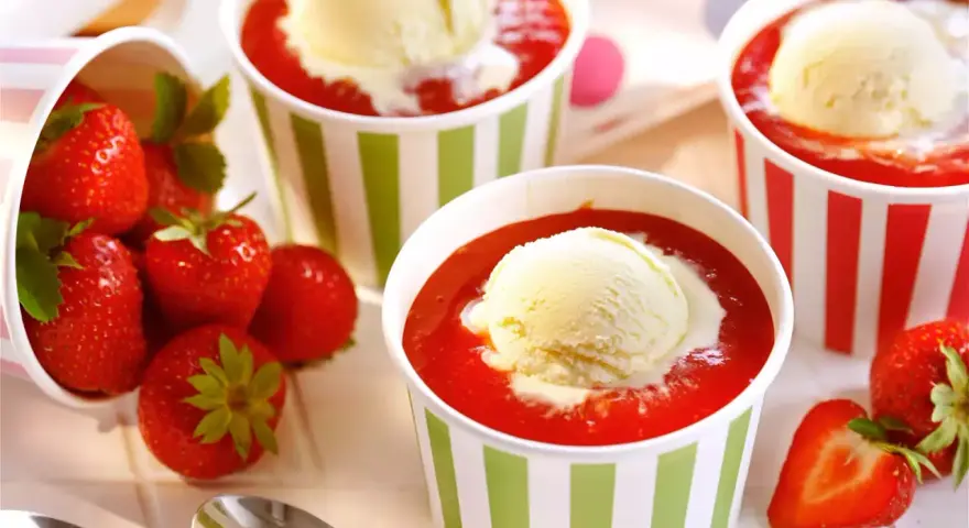Vanilleeis mit Erdbeeren und Sahne in bunten Bechern. Frische Erdbeeren liegen daneben.