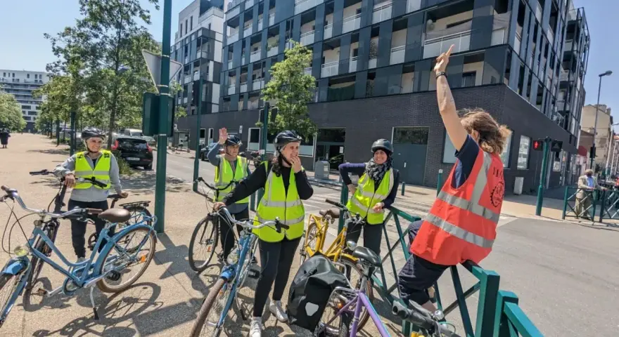 Des adultes souriants, casqués et en gilets fluo, prêts à apprendre à faire du vélo adulte. Ils sont avec leurs vélos, devant un immeuble moderne.