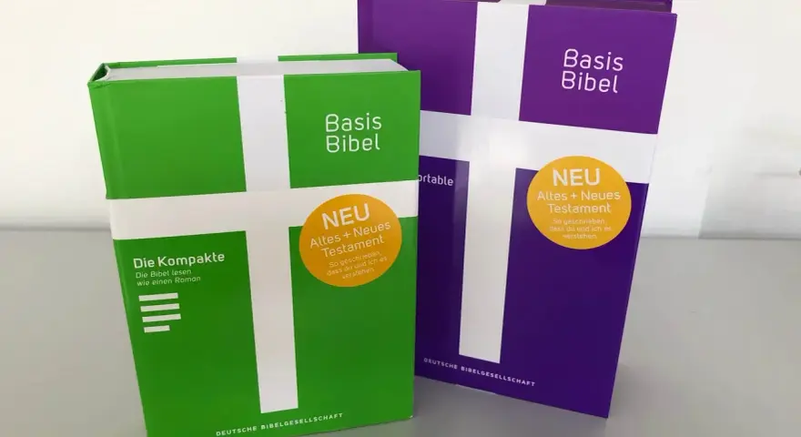Zwei "Basis Bibel" Ausgaben, eine grüne und eine violette, mit dem Aufdruck "NEU Altes + Neues Testament". Diese evangelische Bibel ist so gestaltet, dass sie leicht verständlich ist.