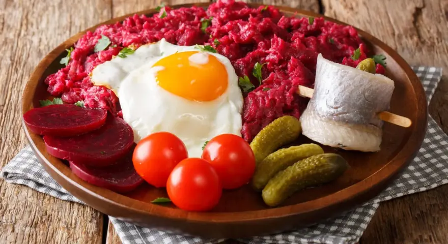 Herzhaftes Labskaus essen: Rote Bete, Spiegelei, Matjes, Cornichons und Tomaten auf einem Holzteller.