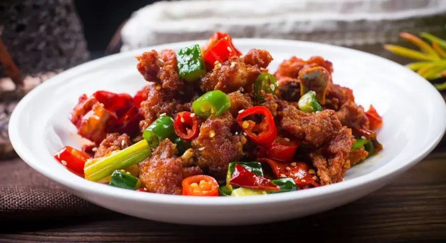 Chrupiący kurczak po kantonsku z papryczkami chili i zieloną cebulką na białym talerzu.