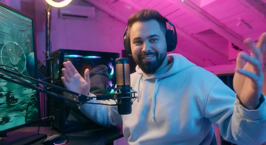 Uśmiechnięty streamer w słuchawkach, przy mikrofonie, tłumaczy, streaming co to jest. W tle komputer i różowe oświetlenie.
