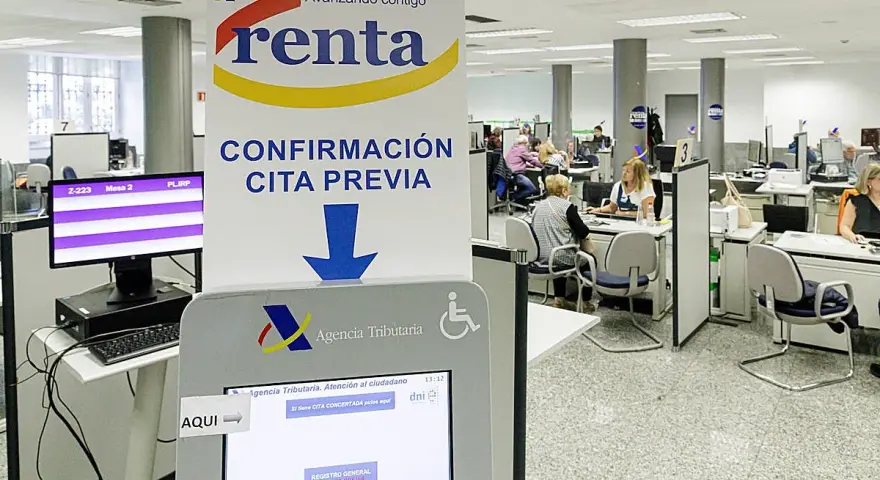 Oficina de la Agencia Tributaria con gente esperando. Un cartel indica "Confirmación cita previa" y una pantalla táctil para gestionar citas, resolviendo la duda de cuanto tarda Hacienda en cobrar la renta.