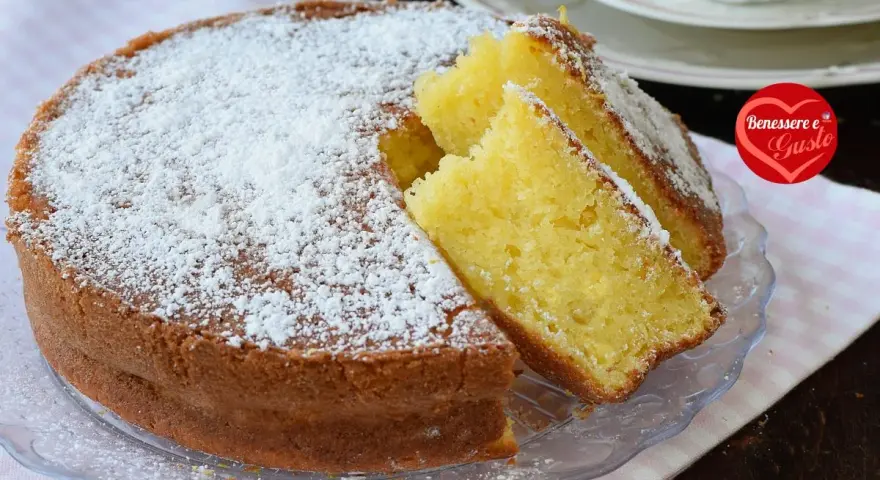 Una soffice torta allo yogurt, cosparsa di zucchero a velo, con due fette tagliate per mostrare la sua morbidezza. Perfetta per la colazione o la merenda.