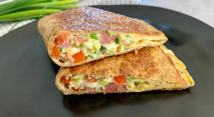 Keto tortilla z jajkiem, warzywami i wędliną, pokrojona na pół, prezentuje apetyczny przekrój.