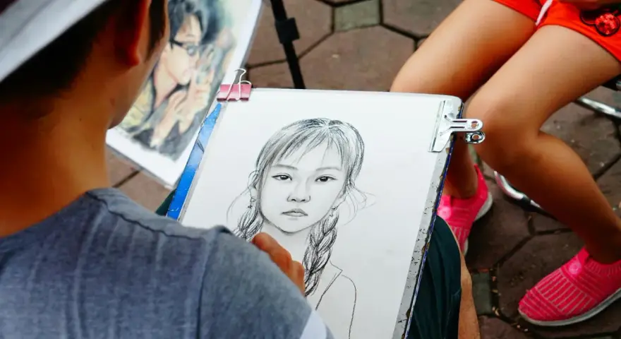 Artysta tworzy portret dziewczynki z warkoczami. Obok leży gotowy rysunek chłopca w okularach.