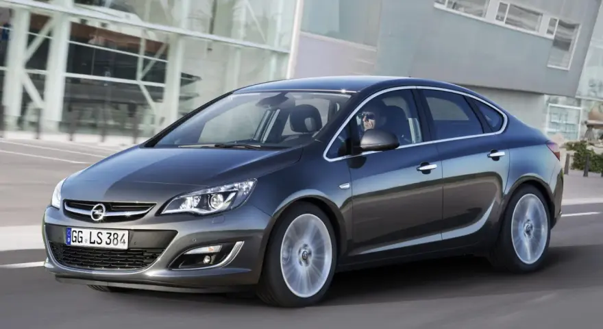 Ciemnoszary Opel Astra J 1.6 benzyna w ruchu, z kierowcą w okularach za kierownicą, na tle nowoczesnego budynku.