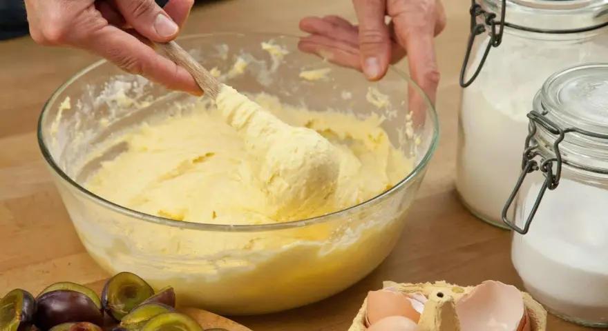 Hände rühren Teig mit Butter oder Margarine in einer Schüssel. Daneben liegen Pflaumen, Eier und Zucker.
