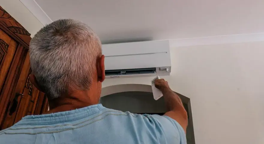 Uomo con telecomando regola il condizionatore per aria fredda, cercando sollievo dall'estate.