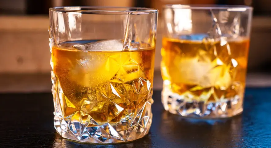 Zwei Gläser mit bernsteinfarbenem on the rocks drink, beleuchtet von warmem Licht.