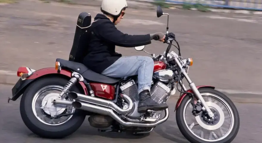 Mężczyzna w kasku jedzie na czerwonym motocyklu Yamaha Virago 250 z futerałem na plecach.