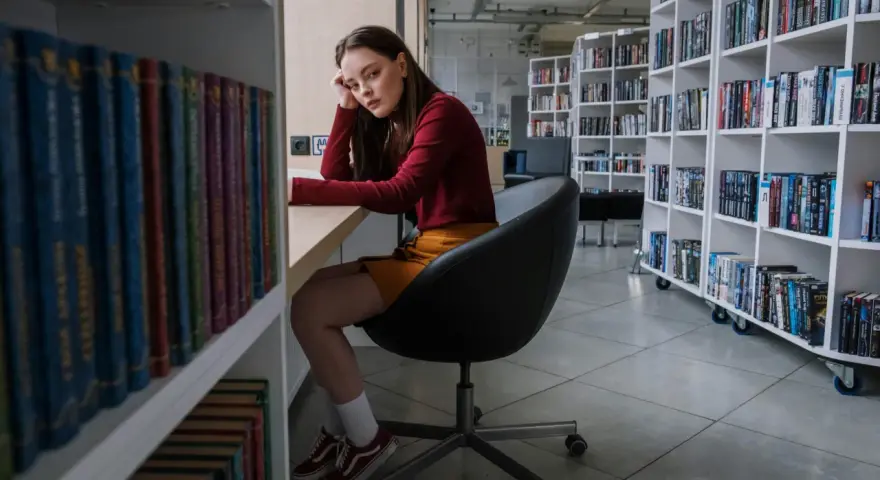 Zamyślona nastolatka w bibliotece, otoczona książkami, odzwierciedla złożoność zachowań nastolatków w okresie dojrzewania.