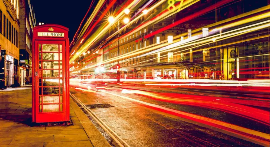 Klasyczna czerwona budka telefoniczna na tle nocnej ulicy, gdzie długi czas naswietlania zdjecia uchwycił smugi świateł samochodowych.
